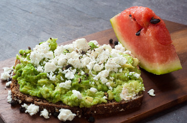 Healthy snack of dark rye bread topped with avocado and crumbled feta cheese served with a slice of fresh water melon.