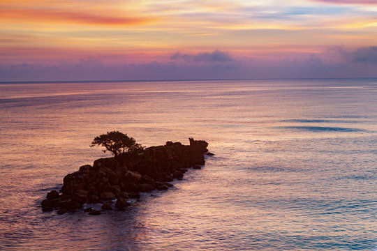 Purple caribbean dawn above a tiny rocky island with a lone tree.