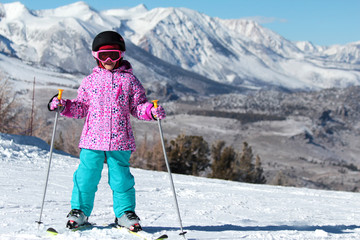 Little Skier girl on a mountain ski. Picture of  little skier girl with  a mountain ski, ski boots, helmet and goggles against the backdrop of snow-capped mountains on an active winter holidays