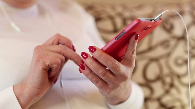 Close up of woman hands with red smartphone. Woman looks and flips through the photos in her smartphone. Businesswoman with red mobile phone with earphones. Female with smartphone