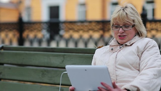 Senior woman sits on the bench near the road and communicates via tablet computer. She is in a merry mood. Woman with Tablet PC sits on the bench and talks on Skype. Pleasant conversation on Skype