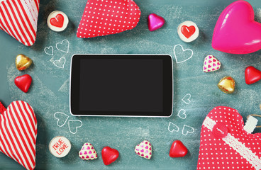top view image of tablet, colorful heart shape chocolates, fabric hearts on blackboard background. valentine's day celebration concept
