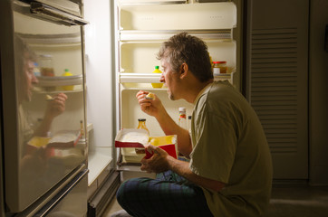A man who has nighttime sleep-related eating disorder sleep eating as he sits in front of a refrigerator eating ice cream out of carton, oblivious to everything around him as he is actually sleeping. - obrazy, fototapety, plakaty