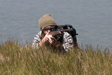 Young hunter with rifle lies on river shore