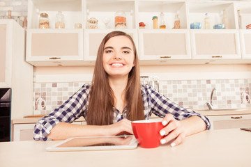 Happy girl sitting in the kitchen with tablet and a cup of coffe