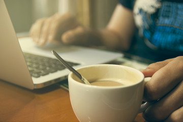 chico moreno con barba tomando el desayuno, un café con leche y trabajando con su ordenador en la cocina de su casa