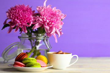 Colorful delicious macaroons on a plate and a Cup of coffee on wooden table on purple background with flowers in a jar of water