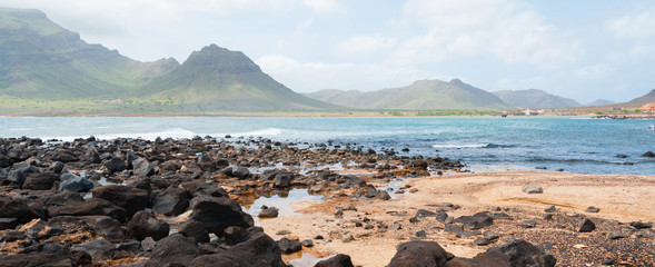 Black rock stone sand beach coast in front of blue sea with mountain background