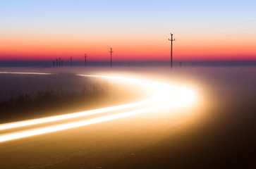 Long Exposure White Car light trails on a road outside at foggy night on blue hour with electrical power lines and pylons disappear over the horizon