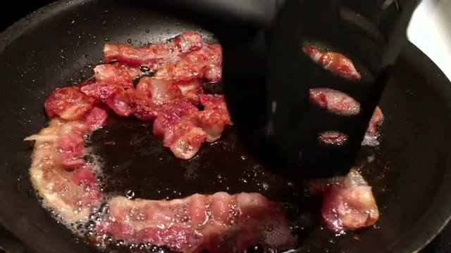 Bacon sizzles in cast iron skillet as black plastic spatula stirs and flips pieces.