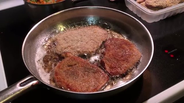 Oil sizzles around breaded meat slices in iron pan on black flat top electric stove with ingredients at edge of shot.