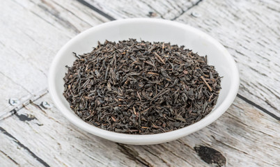 Dried black tea leaves in white bowl over wooden background