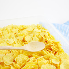 The close up of tasty golden corn flakes and wooden spoon in plastic container box on blue cotton fabric.