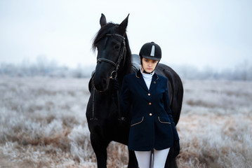 Obraz premium a tall girl with long blond hair in a jockey outfit with a beautiful black horse in an empty snow-covered field in winter