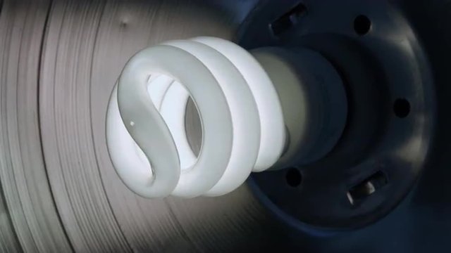 Hand installs a compact fluorescent light bulb into a work light.  Light is barely visible at start of clip and comes on as bulb is fully screwed in.  Big close up with dolly move.