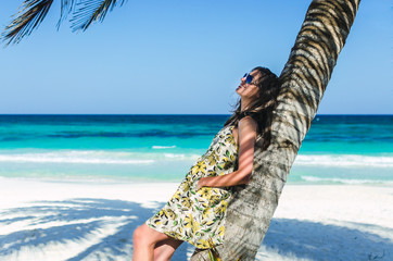 Young adorable caucasian woman in sunglasses and patterned summer dress staying and smiling near palm tree over background of turquoise sea at tropical exotic sandy beach in the Caribbean sea, Mexico