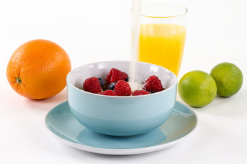 Gesundes Frühstück auf weissem Tisch. Blaue Müslischale mit Müsli, frischen Himbeeren und Heidelbeeren. Auf dem Tsich eine Orange und zwei Limonen. Ein Glas mit Orangensaft im Hintergrund.