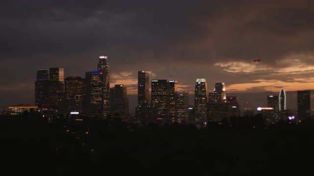 Dusk turns to night over down town Los Angeles, California, and lights come on in the skyscrapers in this establishing shot with a blimp visible, flying over the city.  Originally recorded in 4K