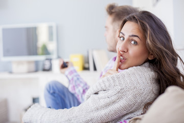 Young couple sitting in the living room watching tv. Woman is making a hush gesture. Silence sign
