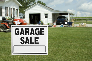 Yard and Garage Sale – A garage sale sign in the front yard of a home. Items for sale in the background. 