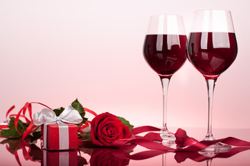 Valentine's decoration with rose, wine glasses, heart and gift boxes. Glossy red table top. Background slightly lit by red.