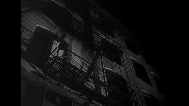 Low angle view of man climbing down fire escape at night, 1940s