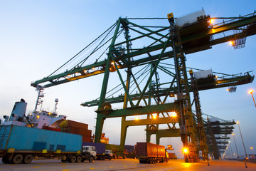 Cranes, cargo containers and trucks at a shipping port during dusk