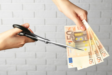 Hands with scissors cutting money on white brick wall background