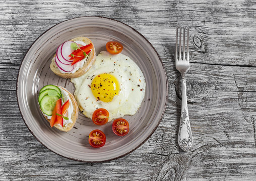 Fried eggs, tomatoes and sandwiches with cucumber, radish and soft cheese. On a light wooden table. Rustic style. Healthy breakfast or snack