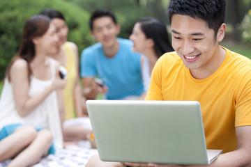 Young man using computer on meadow with friends in background