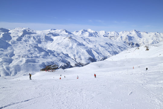 Skiers on ski slopes in high Alps resort, apres ski chalet, with snowy mountain peaks in against blue sky