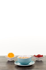 Müsli mit Früchten. Blaue Schale mit Cornflakes. Halbe Orange in weisser eckiger Schale, rote...