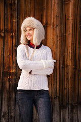 Woman in knitted sweater and furry hat standing near wood wall