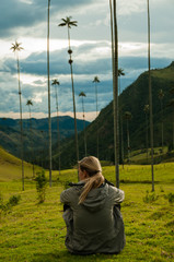 Girl with grey jacket sitting on green grass watching tall palm trees in Cocora Valley