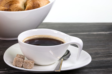 Eine weisse Tasse Kaffee mit drei Würfeln brauner Zucker auf dunklem Holz. Im Hintergrund eine Schale mit Hörnchen.