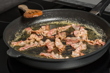 Speck wird in der Pfanne knusprig gebraten. Speck in einer Pfanne mit Fett auf dem Herd. Holzlöffel daneben.