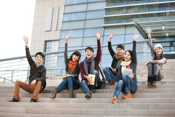 Young college students waving and sitting on steps of university building