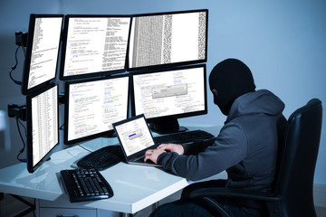 Hacker Using Laptop Against Multiple Monitors At Desk