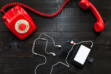 The image shows a hooked off red old retro phone  with a smartphone with white earphones on wooden background
