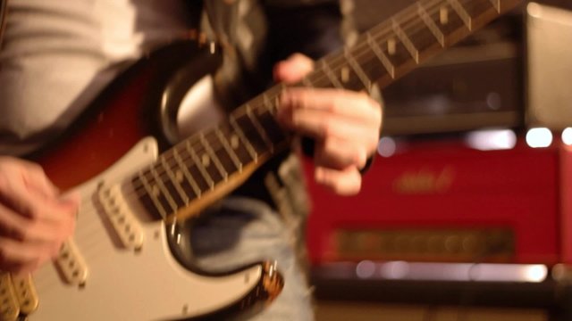 Guitarist. Close-up on a male hand playing hard on a electric guitar. Blurry amplifier & speaker box in background.
