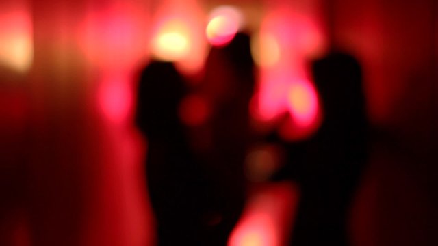 Silhouette of three sexy women dancing in a blurry, red corridor with her hands raised. Medium shot.
