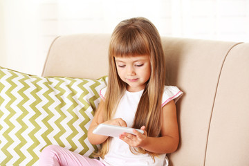 Beautiful little girl  with smart phone sitting on sofa, on home interior background