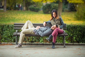 Happy young woman sitting on a bench in a park with her boyfriend lying on her lap