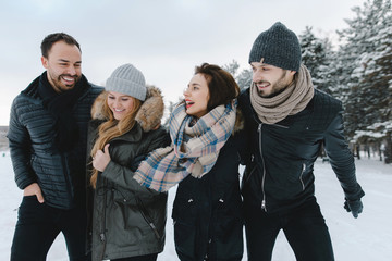 Cute young hipster couples walking in the snow having fun in winter park on a bright day hugging each other and smiling
