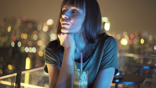 Pensive woman drinking cocktail on terrace in bar at night
