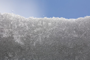 roof skylight window partially covered with melting snow; snow crystals especially in the lower part of image merge and grow larger during melting of snow; blue sky in the background.