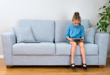 Fototapeta na wymiar Little girl sitting on sofa and using tablet pc. Space for your text.