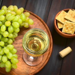 Glass of white wine with white grapes, crackers and a cork, photographed on dark wood with natural light (Selective Focus, Focus on the rim of the wine glass)