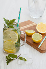 Mojito cocktail with a straw in a jar decorated with fresh mint, wooden board with fresh lemon and lime on it, ice-bucket on the white wooden table. 
