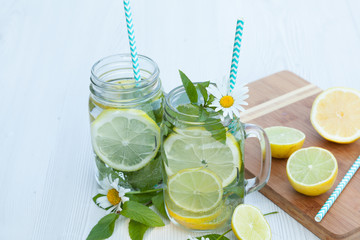 A couple of Mojito cocktails in jars decorated with fresh mint and chamomile flowers, wooden board with fresh lemon and lime on it.